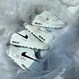 May include: White sneakers with black accents and personalized text. The shoes are arranged on a bed of white fabric. The text includes names and dates, and the brand logo is visible. The overall theme is wedding footwear.