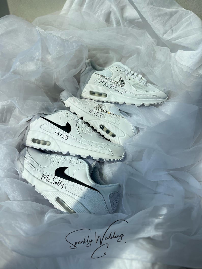 May include: White sneakers with black accents and personalized text. The shoes are arranged on a bed of white fabric. The text includes names and dates, and the brand logo is visible. The overall theme is wedding footwear.