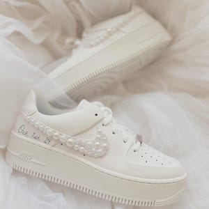 May include: White leather platform sneakers with a pearl embellishment on the swoosh and the text "Oct 1st 24" written on the side.