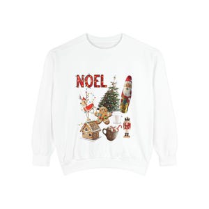 以下が含まれることがあります： お祝いのクリスマスデザインの白いスウェットシャツ。「NOEL」の文字は赤色で表示されています。デザインには、クリスマスツリー、サンタクロース、ジンジャーブレッドマン、トナカイ、その他の休日の要素が含まれています。