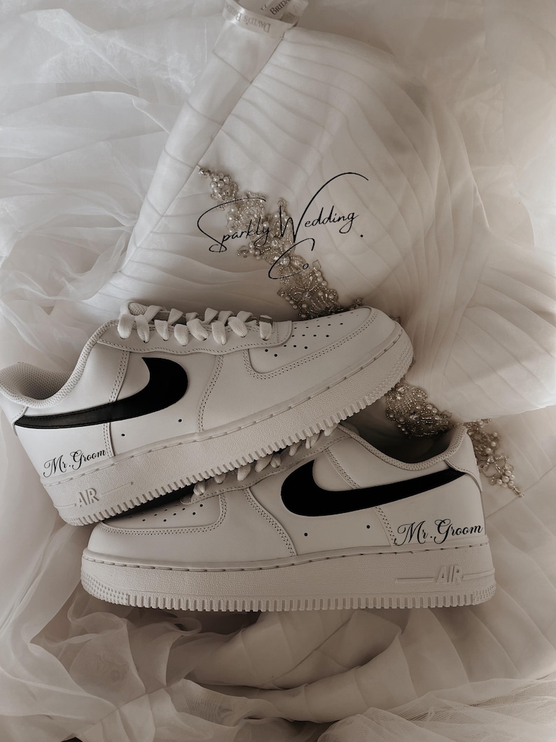 May include: A pair of white sneakers with black swooshes and the text "Mr. Groom" on the side. The sneakers are on a white fabric background.