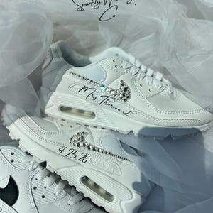 May include: White sneakers with crystal embellishments on the Nike swoosh and accents. The text "Mrs. Thorund" and the date "4.25.26" are written on the shoes. The shoes are part of the "Sparkly Wedding Co." collection.