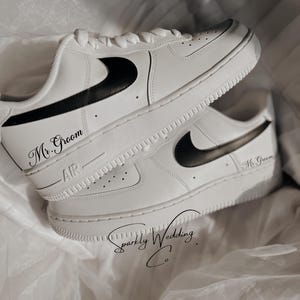 May include: White leather sneakers with black Nike swoosh and the text "Mr. Groom" on the side. The sneakers are on a white fabric background.
