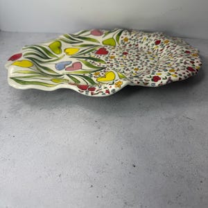 Puede incluir: Un plato decorativo de cer&aacute;mica con borde festoneado. El plato est&aacute; dividido en dos secciones. Un lado presenta hojas verdes y flores coloridas en forma de coraz&oacute;n. El otro lado est&aacute; cubierto con un patr&oacute;n de peque&ntilde;as flores de varios colores.