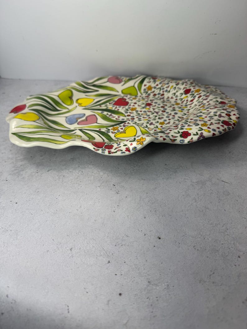 Puede incluir: Un plato decorativo de cer&aacute;mica con borde festoneado. El plato est&aacute; dividido en dos secciones. Un lado presenta hojas verdes y flores coloridas en forma de coraz&oacute;n. El otro lado est&aacute; cubierto con un patr&oacute;n de peque&ntilde;as flores de varios colores.