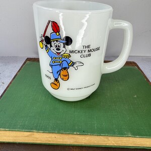 Peut inclure: Tasse en c&eacute;ramique blanche avec Mickey Mouse en uniforme de fanfare bleu, tenant une baguette. Le texte "THE MICKEY MOUSE CLUB" et "1955" est visible. Un objet de collection vintage de Walt Disney Productions.