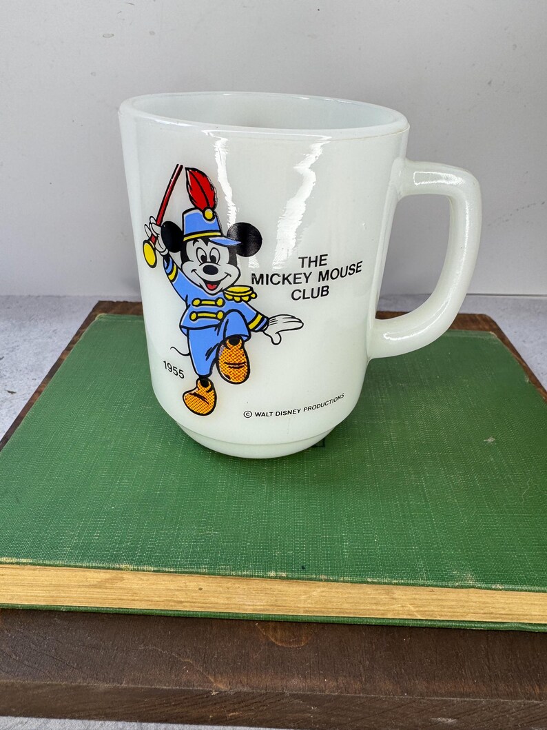 Peut inclure: Tasse en c&eacute;ramique blanche avec Mickey Mouse en uniforme de fanfare bleu, tenant une baguette. Le texte "THE MICKEY MOUSE CLUB" et "1955" est visible. Un objet de collection vintage de Walt Disney Productions.