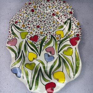 Puede incluir: Una fuente de cer&aacute;mica blanca con un dise&ntilde;o floral. La fuente tiene un borde festoneado y est&aacute; decorada con flores coloridas en forma de coraz&oacute;n en rojo, amarillo, rosa y azul, con hojas verdes.
