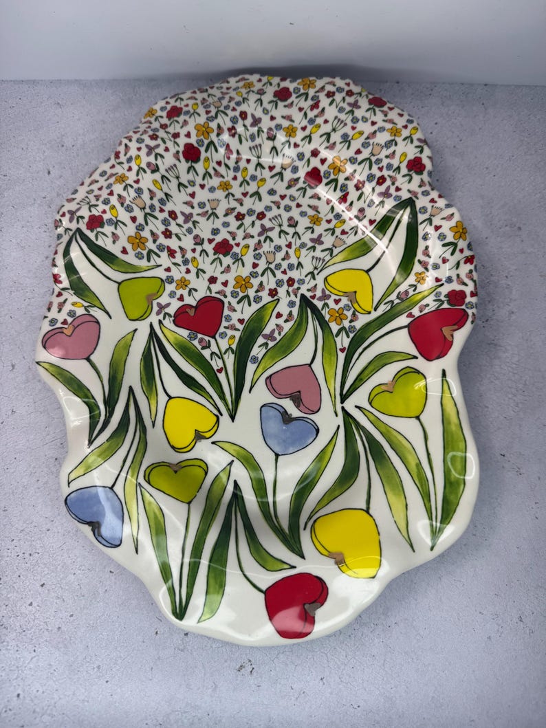 Puede incluir: Una fuente de cer&aacute;mica blanca con un dise&ntilde;o floral. La fuente tiene un borde festoneado y est&aacute; decorada con flores coloridas en forma de coraz&oacute;n en rojo, amarillo, rosa y azul, con hojas verdes.