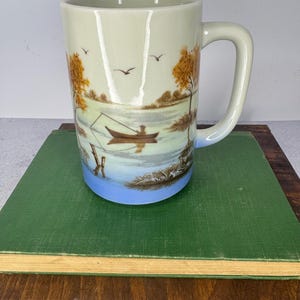 May include: A tall ceramic mug with a glossy finish featuring a painted landscape scene of a person in a small boat on a lake. The design includes golden-brown trees, flying birds, and a blue base. The mug has a curved handle on the right side.