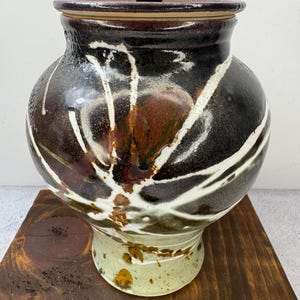 May include: A handcrafted ceramic jar with a lid. The jar features a dark brown and black glaze with white, abstract, linear designs. The base is a light green color with brown speckles. The lid has a dark brown knob.