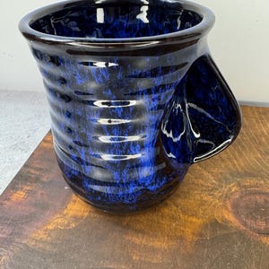 May include: A ceramic mug with a unique handle. The mug features a dark blue and black speckled glaze with horizontal ridges. The interior and rim are a darker shade of blue. The mug sits on a wooden surface.
