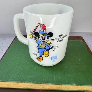 Peut inclure: Tasse en c&eacute;ramique blanche avec une illustration color&eacute;e de Mickey Mouse en uniforme bleu, tenant un b&acirc;ton. La tasse porte l'inscription "The Mickey Mouse Club" et "1955". Un logo Pepsi est &eacute;galement visible.