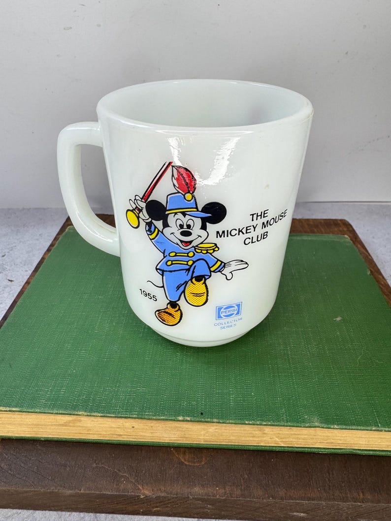 Peut inclure: Tasse en c&eacute;ramique blanche avec une illustration color&eacute;e de Mickey Mouse en uniforme bleu, tenant un b&acirc;ton. La tasse porte l'inscription "The Mickey Mouse Club" et "1955". Un logo Pepsi est &eacute;galement visible.