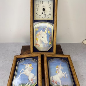 May include: A rectangular wall clock with a light oak wood frame, featuring a white clock face with black numerals and the Elgin brand name. Below the clock face is a decorative panel depicting a white unicorn with a golden horn and floral accents.