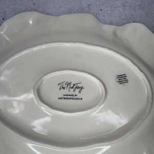Puede incluir: Plato para servir de cer&aacute;mica blanquecina con borde ondulado. El plato tiene un centro ovalado con el texto "The Mud Fairy" y "exclusively for ANTHROPOLOGIE". La parte posterior del plato tiene una etiqueta "Handwash Only".
