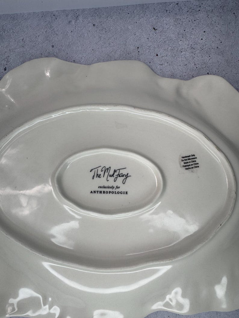 Puede incluir: Plato para servir de cer&aacute;mica blanquecina con borde ondulado. El plato tiene un centro ovalado con el texto "The Mud Fairy" y "exclusively for ANTHROPOLOGIE". La parte posterior del plato tiene una etiqueta "Handwash Only".