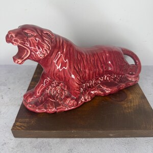 May include: A ceramic tiger figurine in a glossy burgundy finish, posed in a roaring position. The sculpture is placed on a dark brown wooden base, showcasing the detailed craftsmanship of the animal figure.