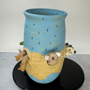 May include: A handcrafted ceramic vase with a light blue exterior and a whimsical design. The vase features a koala and panda bear, with a tan-colored boat-like structure. The vase is on a black stand.