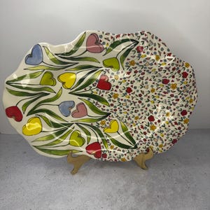 Puede incluir: Una fuente decorativa de cer&aacute;mica con borde festoneado. Un lado presenta flores en forma de coraz&oacute;n en rojo, amarillo, azul y rosa con tallos verdes. El otro lado tiene un estampado floral con peque&ntilde;as flores de varios colores.