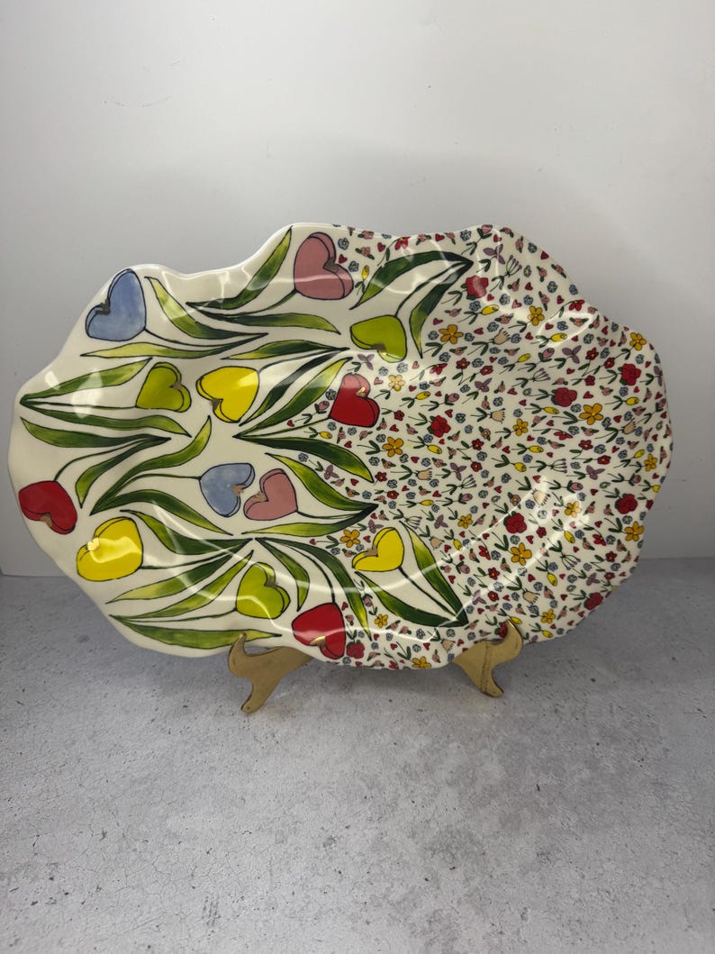 Puede incluir: Una fuente decorativa de cer&aacute;mica con borde festoneado. Un lado presenta flores en forma de coraz&oacute;n en rojo, amarillo, azul y rosa con tallos verdes. El otro lado tiene un estampado floral con peque&ntilde;as flores de varios colores.