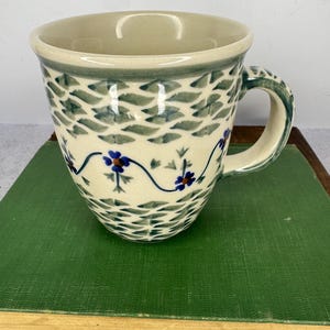 May include: Cream-colored ceramic mug with a green and blue floral design. The mug features a curved handle and a wide rim. The exterior is decorated with a repeating pattern of green leaves and blue flowers.