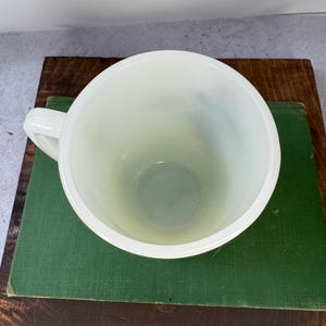 Peut inclure: Mug cr&egrave;me avec une anse, vue de dessus. Le mug est pos&eacute; sur un livre vert, lui-m&ecirc;me pos&eacute; sur une surface en bois. L'int&eacute;rieur du mug est visible, et l'esth&eacute;tique g&eacute;n&eacute;rale est vintage.