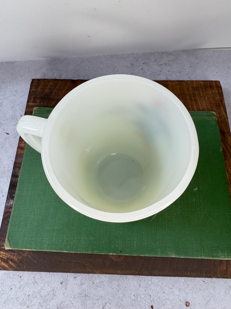 Peut inclure: Mug cr&egrave;me avec une anse, vue de dessus. Le mug est pos&eacute; sur un livre vert, lui-m&ecirc;me pos&eacute; sur une surface en bois. L'int&eacute;rieur du mug est visible, et l'esth&eacute;tique g&eacute;n&eacute;rale est vintage.