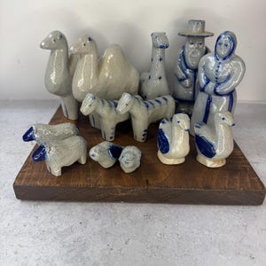 May include: A collection of glazed ceramic figurines featuring two camels, two horses, two sheep, two small animals, two geese, and two human figures. The pieces are primarily light gray with blue painted details and accents throughout the set.