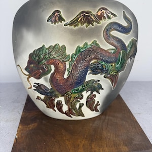 May include: A ceramic vase with a gray background and a detailed dragon design in shades of red, green, and blue. The dragon is the focal point, with mountain-like shapes above it. The vase sits on a wooden base.