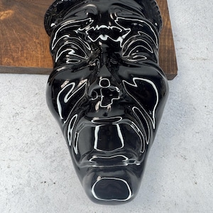 May include: A glossy black ceramic mask with white accent lines. The mask has a detailed face with a prominent nose, mouth, and eyes. The mask is mounted on a wooden surface.