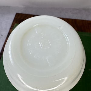 Peut inclure: Une tasse en verre blanc opaque avec une anse. Le fond de la tasse porte du texte en relief et un logo carr&eacute;. La tasse est pos&eacute;e sur une surface verte.