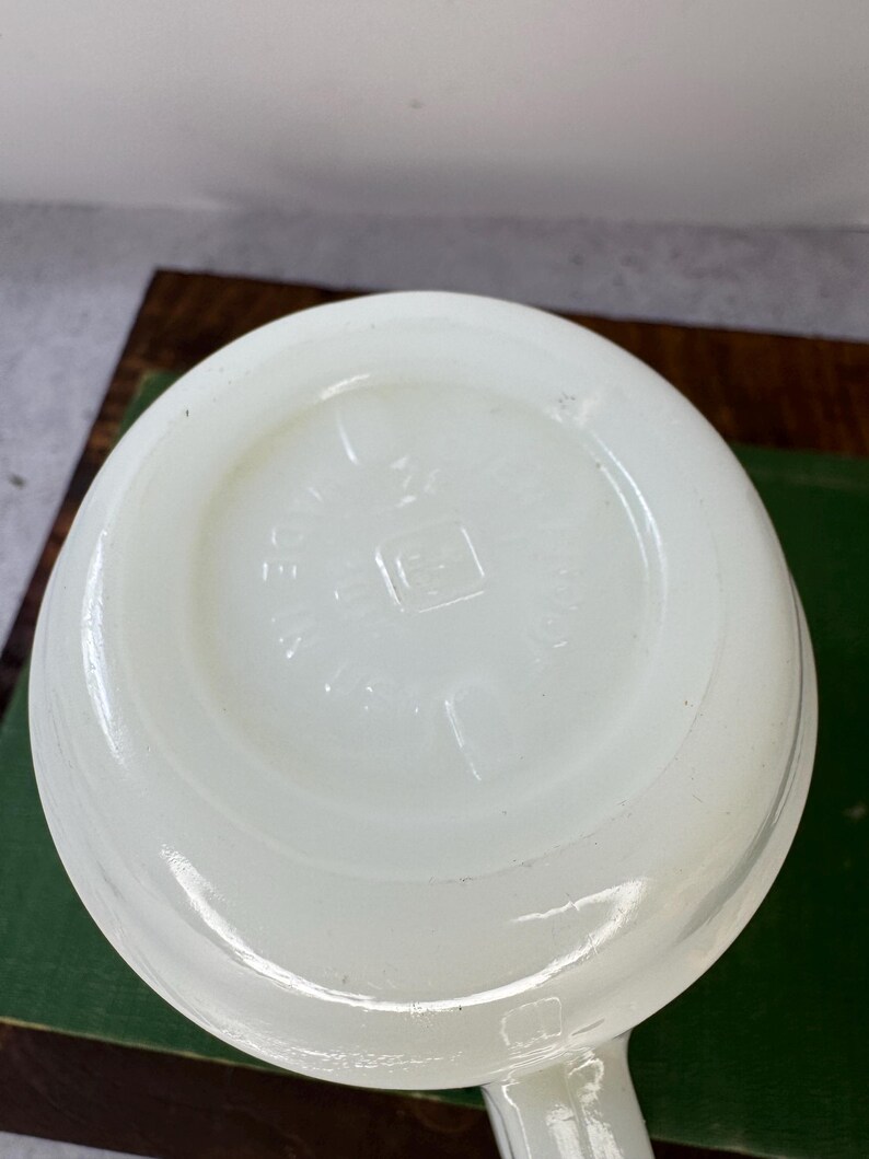 Peut inclure: Une tasse en verre blanc opaque avec une anse. Le fond de la tasse porte du texte en relief et un logo carr&eacute;. La tasse est pos&eacute;e sur une surface verte.