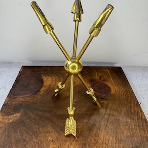 May include: A brass arrow-themed decorative object. The object features three arrows radiating from a central sphere, with a fourth arrow pointing downward. The arrows have a gold finish and are arranged on a wooden surface.