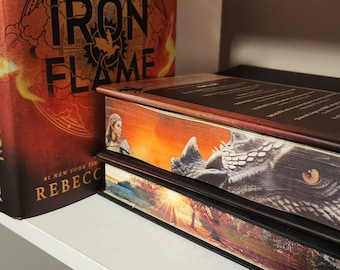 Puede incluir: Una estantería con tres libros apilados uno encima del otro. El libro de arriba tiene el título "Iron Flame" y tiene una portada roja, naranja y amarilla con un diseño de dragón. El libro del medio tiene una portada marrón con un dragón y una mujer en la portada. El libro de abajo tiene una portada marrón con una escena de bosque.