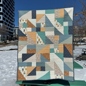 May include: A quilt with a geometric pattern in shades of blue, brown, and beige. The quilt is made up of squares and triangles, and it has a modern, abstract design.