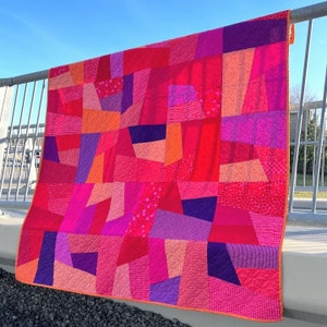 May include: A patchwork quilt made of various shades of pink and purple fabric. The quilt is made up of many different geometric shapes, creating a modern and abstract design.