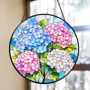 May include: A circular stained glass suncatcher featuring clusters of blue, pink, and white hydrangea flowers with green leaves. The design is enclosed in a black metal frame and hangs from a metal chain, showcasing varied glass textures.