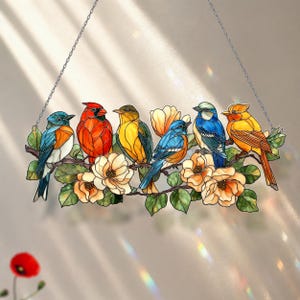 May include: A stained glass suncatcher featuring six colorful birds perched on a flowering branch. The birds are rendered in shades of blue, red, yellow, and orange, surrounded by light peach-colored blossoms and green leaves, suspended by a silver chain.