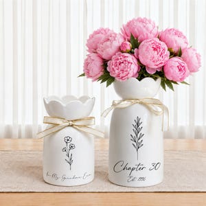 May include: Two white ceramic vases with scalloped rims and gold ribbon bows. The smaller vase features a floral line drawing and the text In My Grandma Era. The larger vase holds pink peonies and displays a leaf drawing with Chapter 30 Est. 1996.