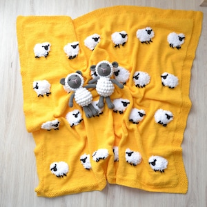 Op de afbeelding: Een gele gebreide babydeken met een wit schapenontwerp. Twee kleine grijze en witte gehaakte schaapjes liggen op de deken.