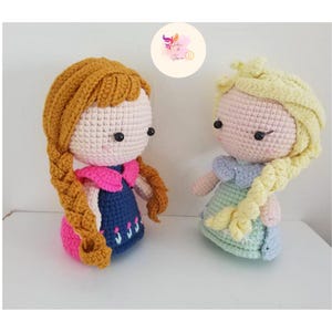 May include: Two crocheted dolls, one with brown hair and a blue dress, the other with blonde hair and a light green dress.