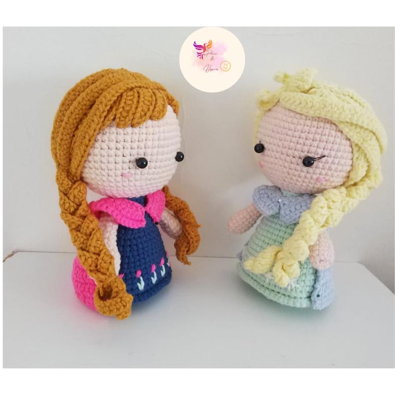 May include: Two crocheted dolls, one with brown hair and a blue dress, the other with blonde hair and a light green dress.
