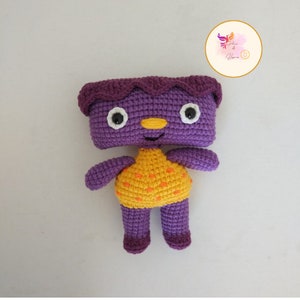 Puede incluir: Juguete de crochet morado con un vestido de lunares amarillos y naranjas. El juguete tiene una cara sonriente con ojos blancos y una nariz amarilla.