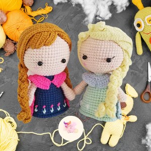 May include: Two crocheted dolls, one with brown hair and a blue dress, the other with blonde hair and a green dress. The dolls are holding hands and are standing on a gray surface.