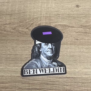 May include: A sticker featuring a black and white image of Benjamin Franklin wearing a welding mask. The text "BEN WELDIN" is printed in white block letters below the image. The welding mask has a purple lens.
