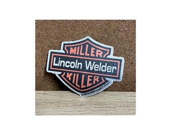 May include: A sticker with a shield-shaped design featuring the words "MILLER," "Lincoln Welder," and "KILLER" in a bold, stylized font. The sticker has a black background with white and orange accents. The website address "@WeldShop21.com" is at the bottom.