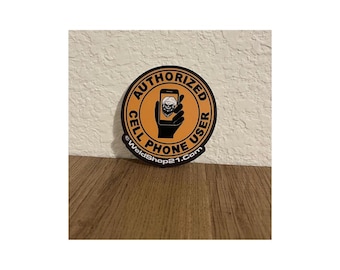 May include: A round sticker with an orange background and black border. The sticker reads "AUTHORIZED CELL PHONE USER" in a circular pattern. A black hand holds a phone with a skull graphic. The website "WeldShop21.com" is at the bottom.