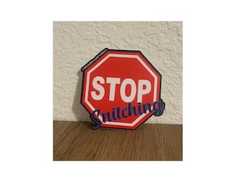May include: A red, white, and black octagon-shaped sticker with the words "STOP Snitching" in white and blue lettering. The sticker is placed on a wooden surface.