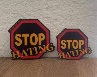 May include: Two octagonal signs with the words "STOP HATING". The signs are black with yellow text and a red border. The smaller sign is to the right of the larger sign. The website address "Weldshop21.com" is printed on the top left corner of each sign.